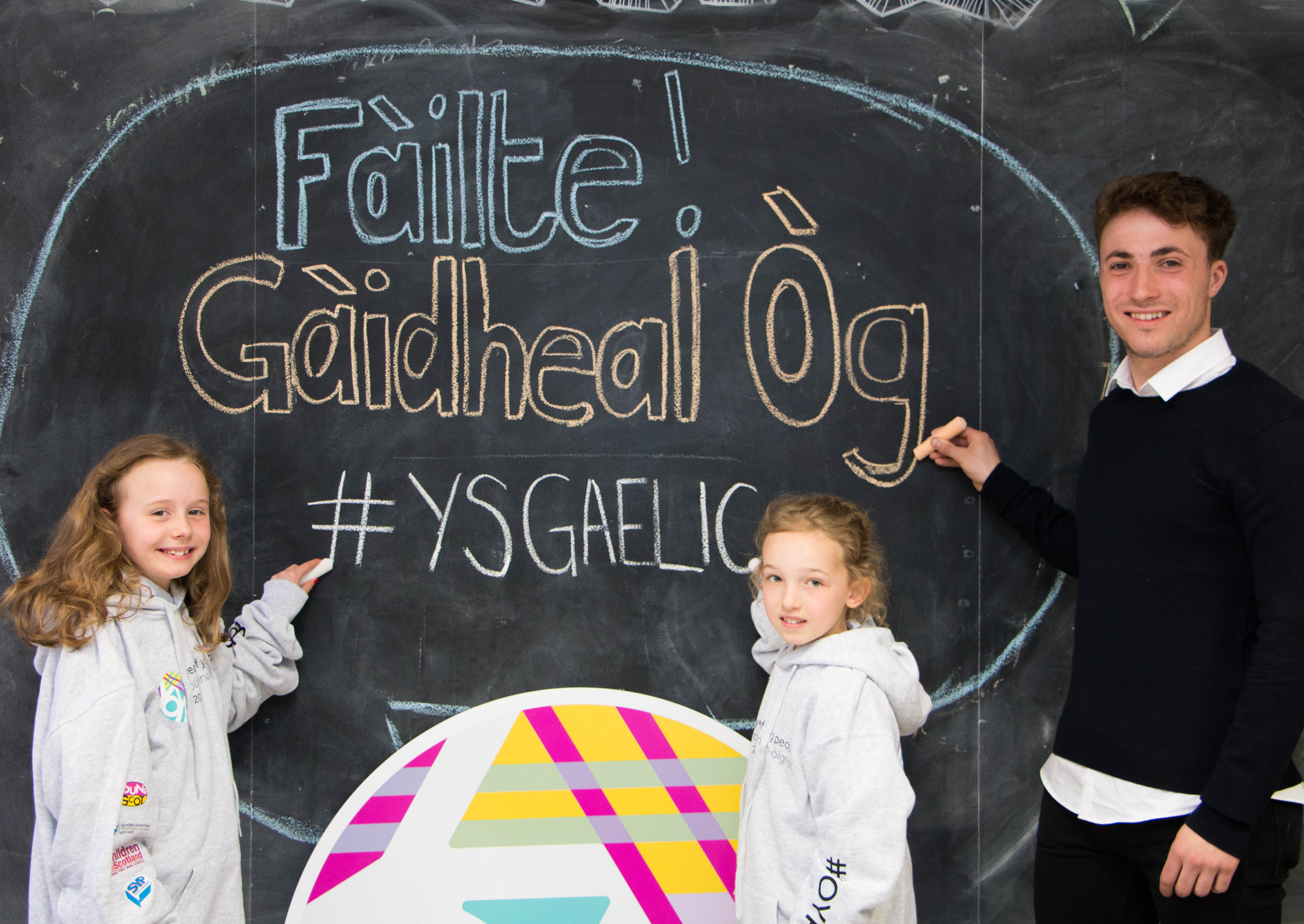 Picture: The three school children holding chalk and writing  "Fàilte! Gàidheal Òg! #YSGaelic "Welcome! Young geals" on the black chalkboard
