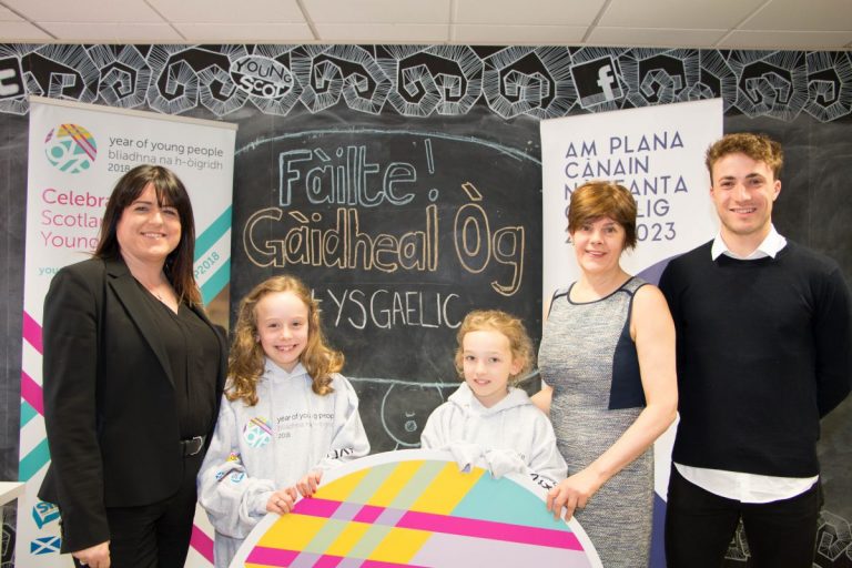Picture: Louise MacDonald, CEO of Young Scot, Shona MacLennan, CEO of Bòrd na Gàidhlig, one secondary-school age and two primary school age children standing in front of a large chalkboard. A National Gaelic Language Plan banner and a Young Scot Year of Young People banner are behind them.
