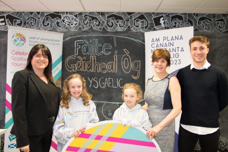 Picture: Louise MacDonald, CEO of Young Scot, Shona MacLennan, CEO of Bòrd na Gàidhlig, one secondary-school age and two primary school age children standing in front of a large chalkboard. A National Gaelic Language Plan banner and a Young Scot Year of Young People banner are behind them.