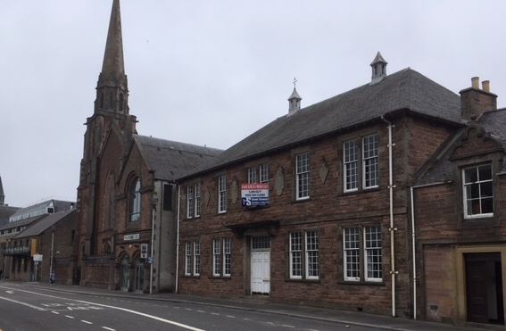 Picture: Red stone local heritage building with a white door and a for sale sign on one of 16 sash windows. It is next door to a red stone church and on a city road. 