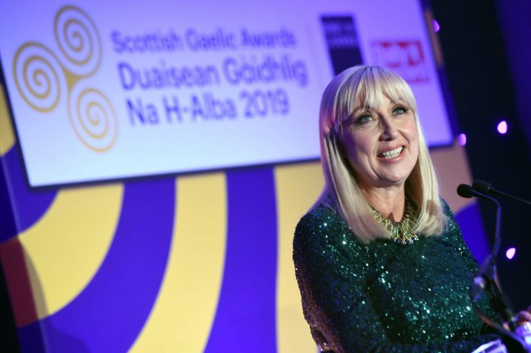 Picture: A female presenter speaking into a microphone on stage with a large sign behind that reads "Scottish Gaelic Awards / Duaisean Gàidhlig na h-Alba 2019"