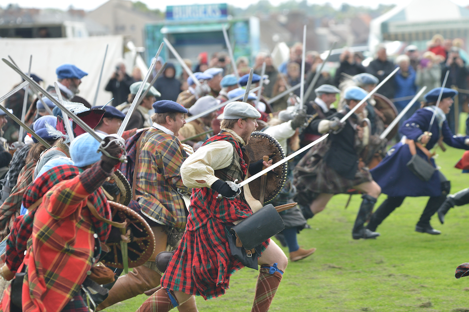 Picture: A crowd of men reenacting a battle, all wearing kilts and tweed flatcaps and charging forwards in a field with shields and swords
