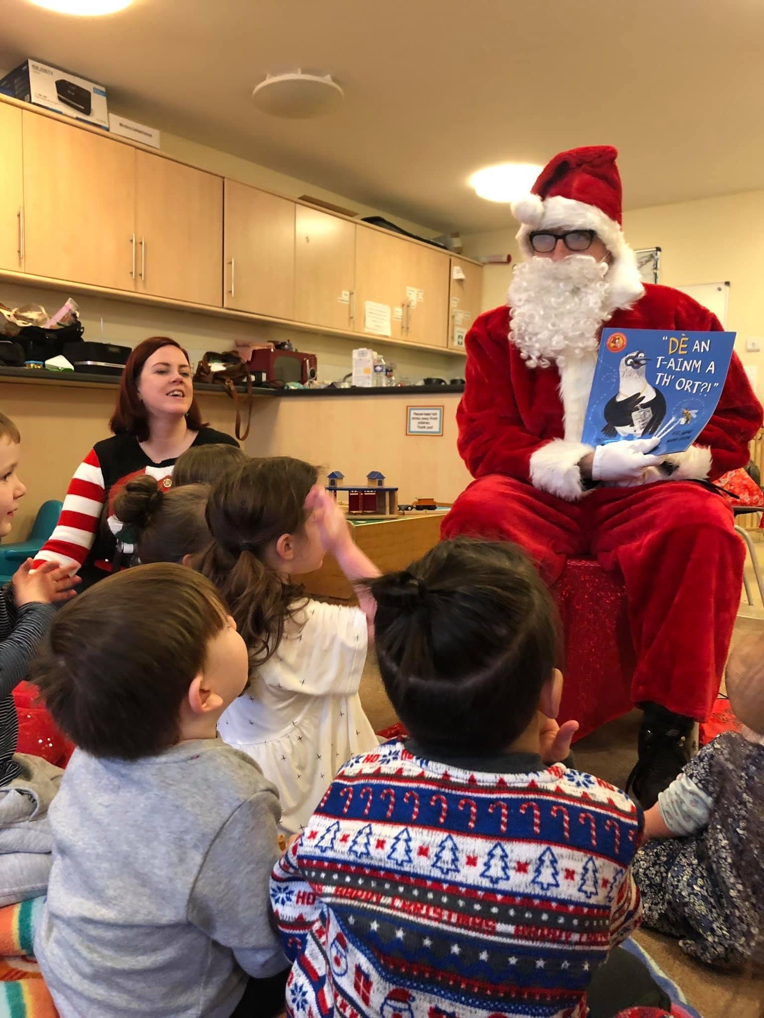 Picture: Santa claus reading "Dè an t-ainm a th'ort?" to children and staff member.