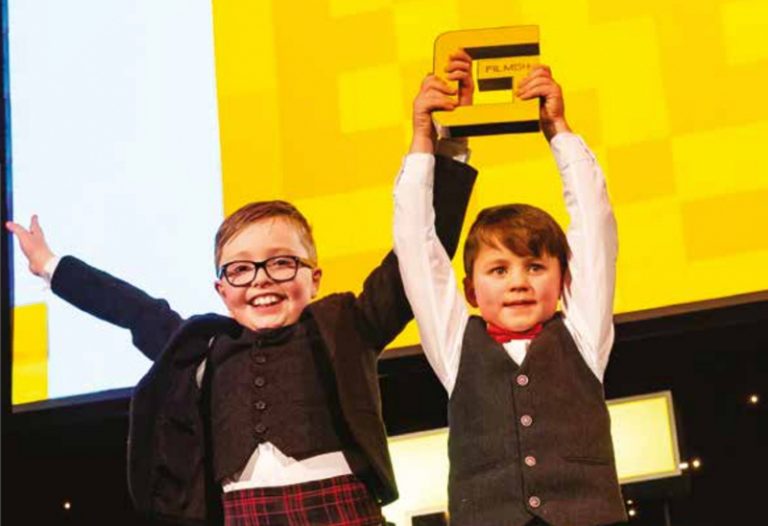 Picture: Two young boys in suits holding a FilmG award they have just won high in the air on the stage at the FilmG awards.